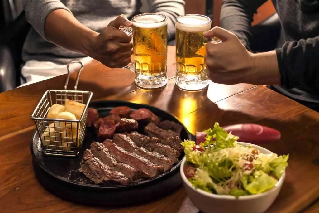 Foto com uma grelha com churrasco em cima e acompanhamentos ao lado, e ao fundo é possível ver duas mãos segurando copos com cerveja.