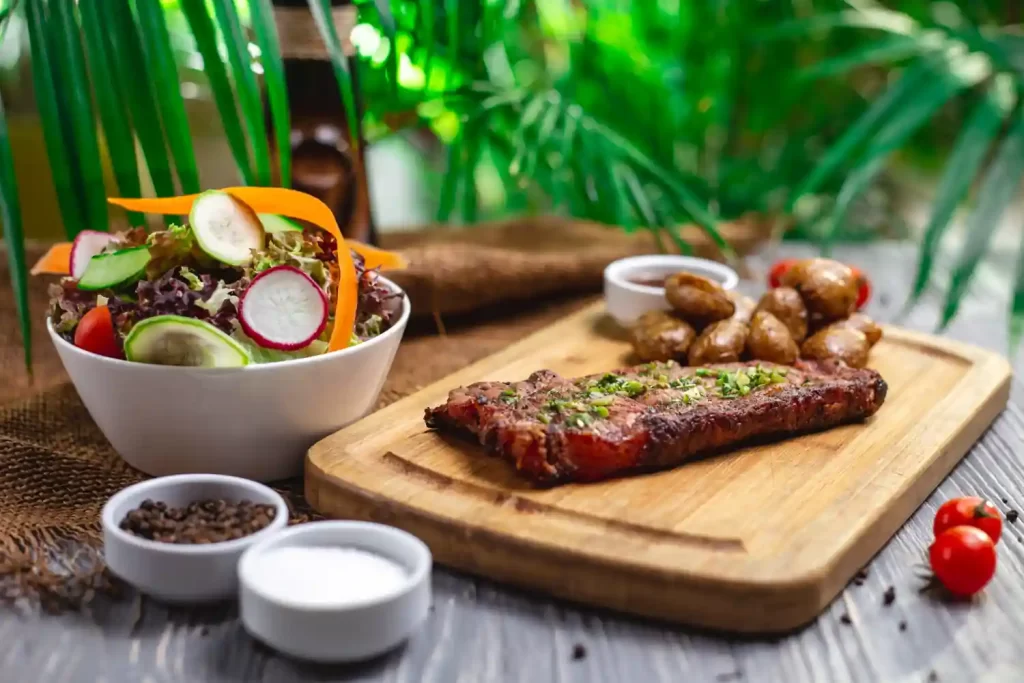 Imagem com uma carne grelhada no centro da mesa junto com alguns acompanhamento ao lado, como por exemplo uma salada e algumas batatas.